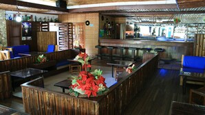 A bar area with wooden furniture, a central bar counter, and a seating area with blue cushioned chairs. - Hotel Guadaira Resort (Melgar)