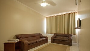 Een woonkamer met een bruine leren bank, een bijpassende fauteuil, een houten bijzettafeltje en een groot raam met beige gordijnen.