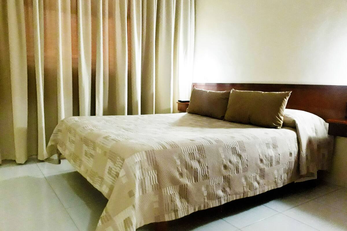 Una cama bien hecha con una colcha estampada y dos almohadas, un cabecero de madera y una ventana con cortinas.