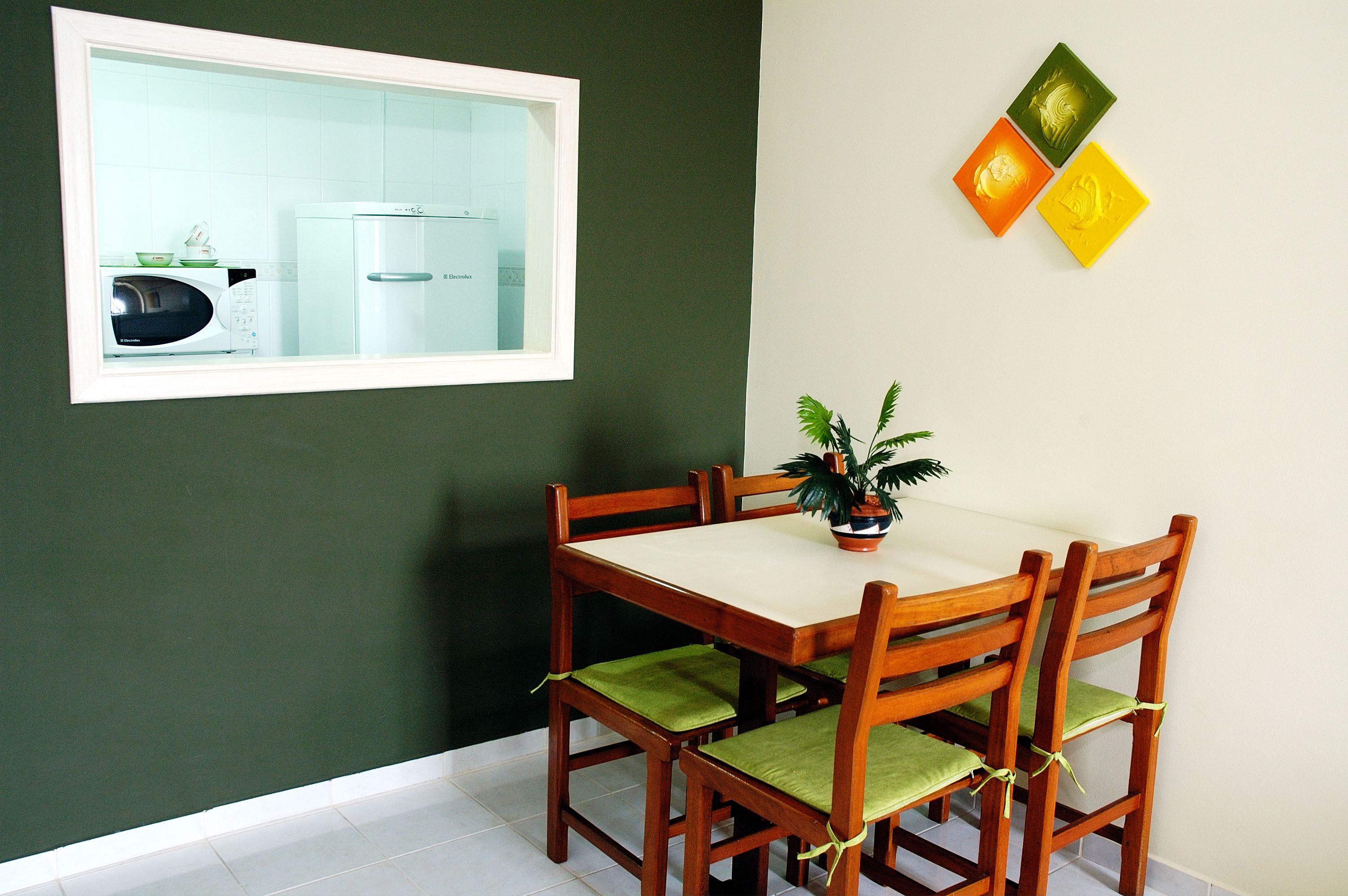 Uma área de refeições com mesa e cadeiras, uma planta em vaso e um micro-ondas visíveis através de uma janela.