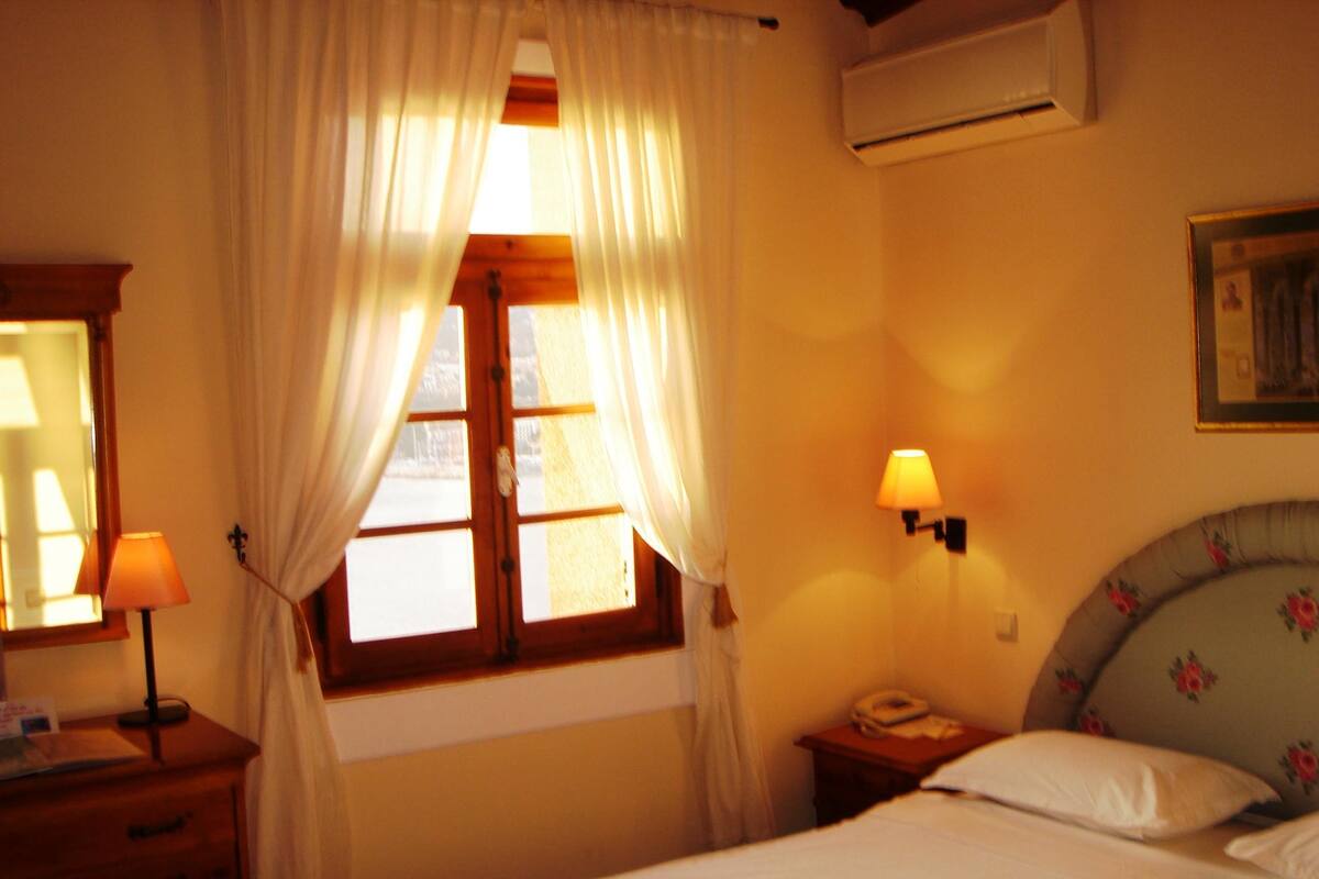 A bedroom with a bed, a nightstand, a lamp, a window with curtains, and an air conditioning unit.