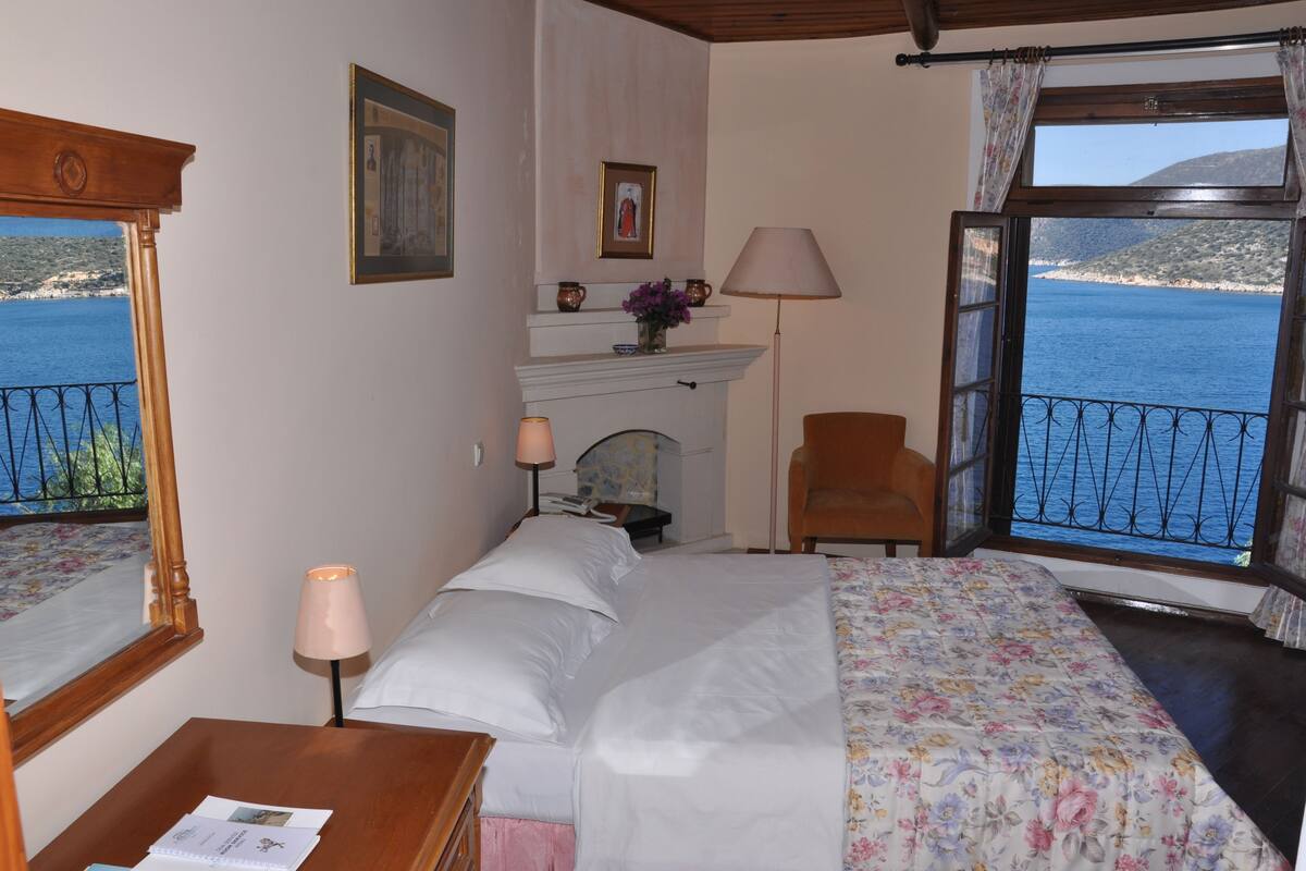A bedroom with a bed, a nightstand, a chair, a fireplace, and a view of the sea.
