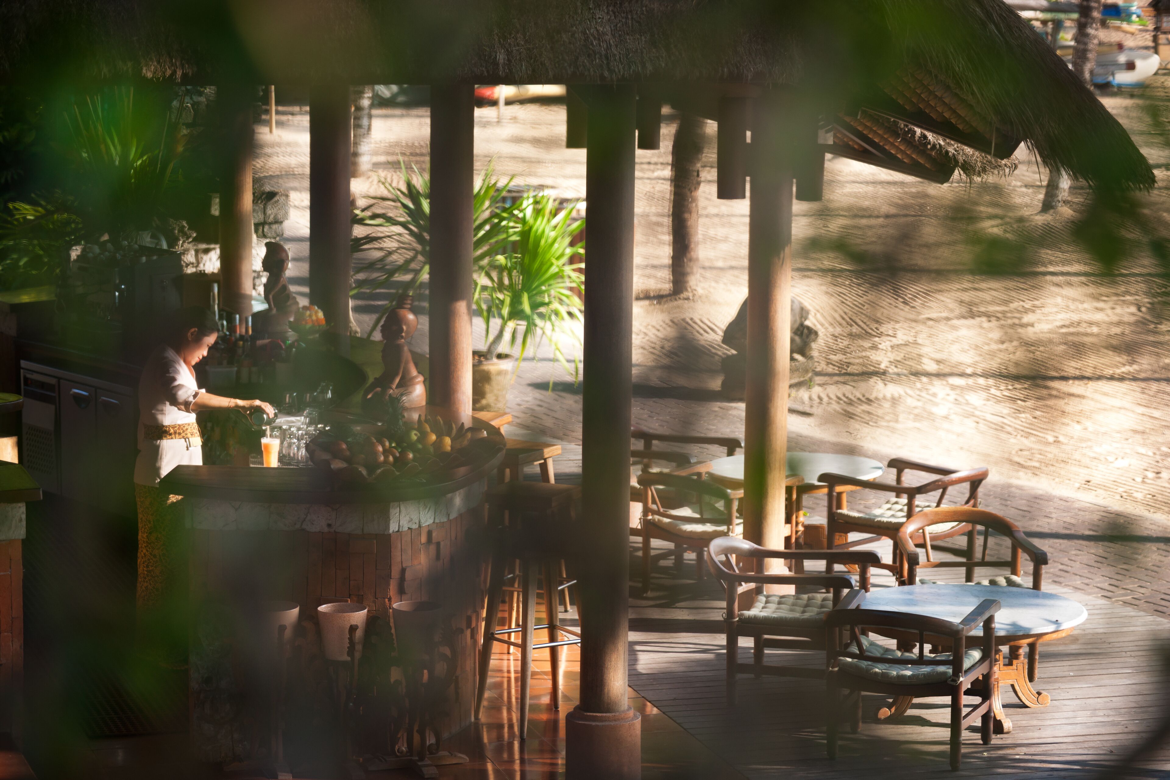 Une terrasse en bois avec des sièges d’extérieur, comprenant des chaises et des tables, entourée de palmiers et surmontée d’une structure à toit de chaume.