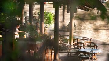 A wooden deck with outdoor seating, including chairs and tables, surrounded by palm trees and a thatched roof structure.