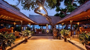 An outdoor covered area with thatched roofs, potted plants, and a central statue.