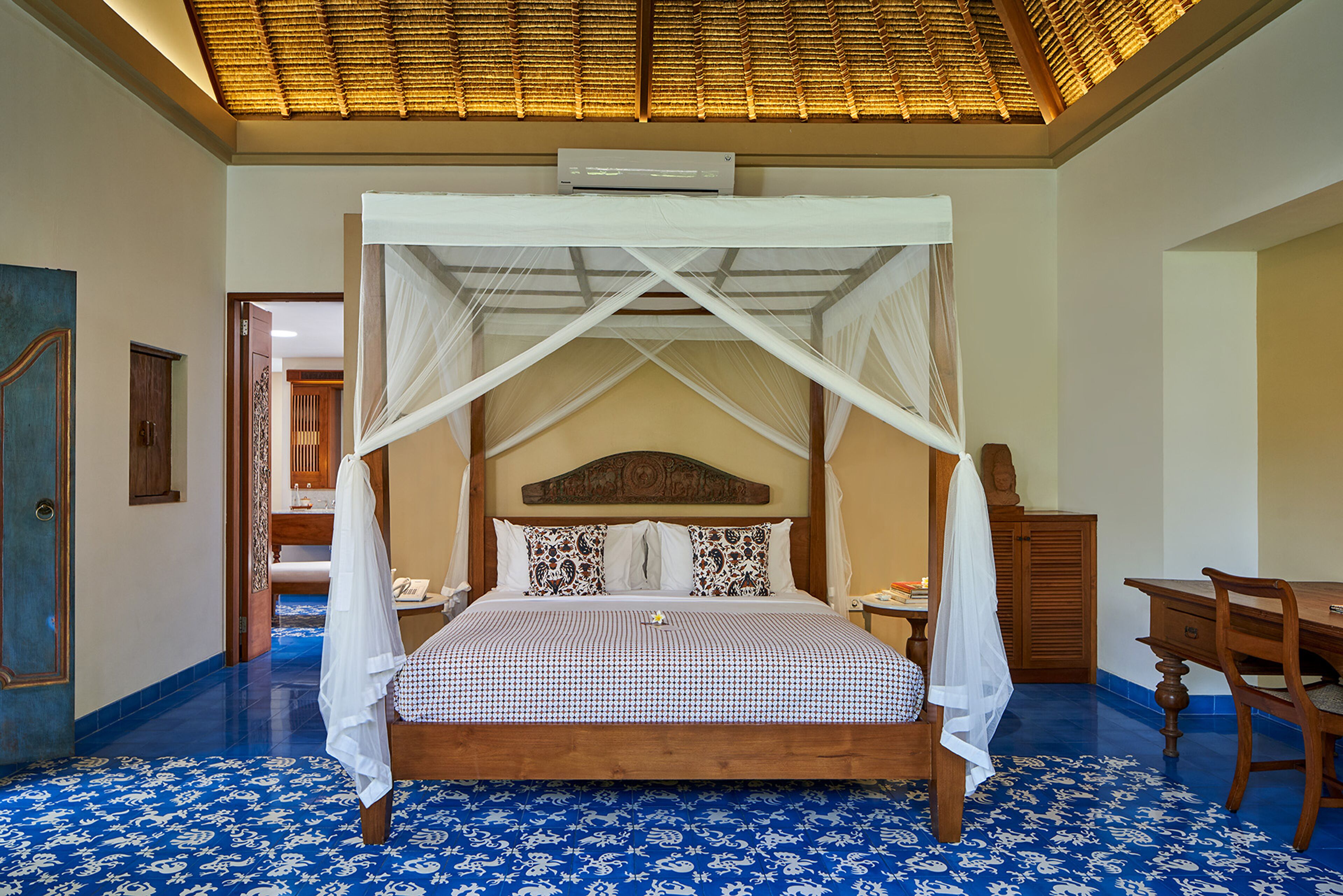A four-poster bed with a canopy, a wooden desk, and a chair in a room with a blue patterned floor and a thatched ceiling.