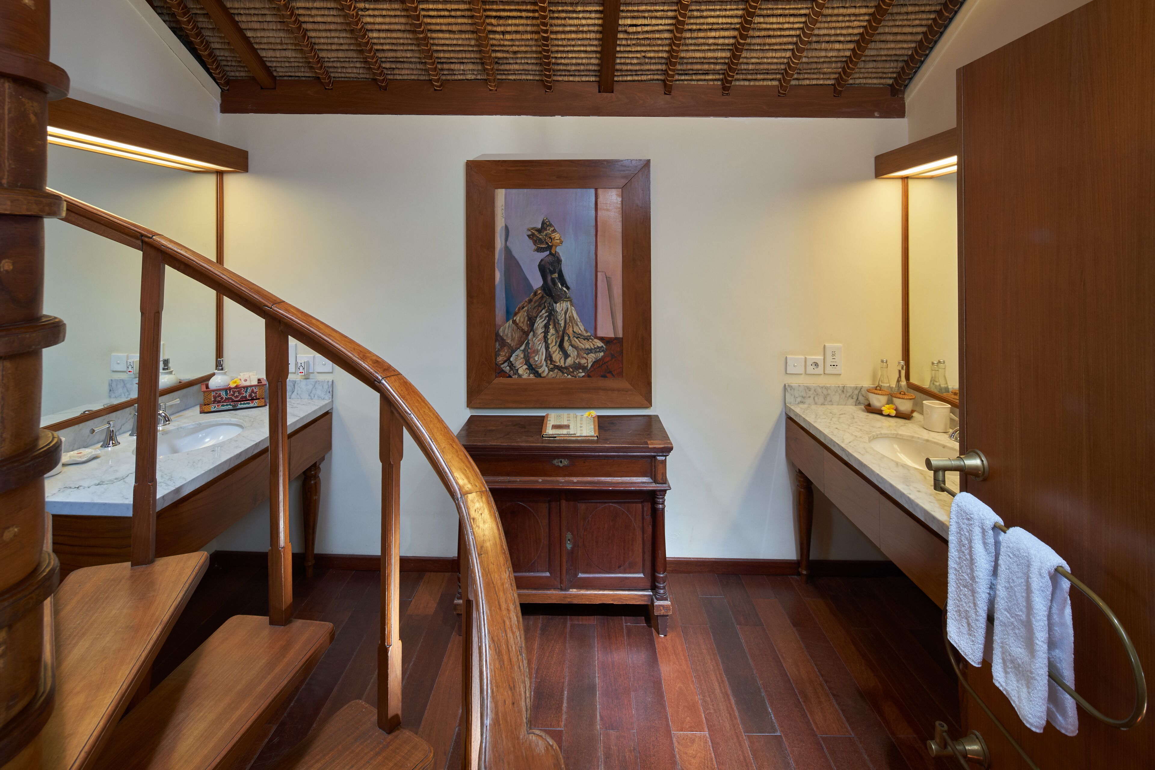 Una escalera de madera conduce a un baño con lavamanos de encimera de mármol, tocador de madera y un cuadro enmarcado sobre el espejo.