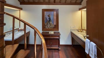 Una escalera de madera conduce a un baño con lavamanos de encimera de mármol, tocador de madera y un cuadro enmarcado sobre el espejo.