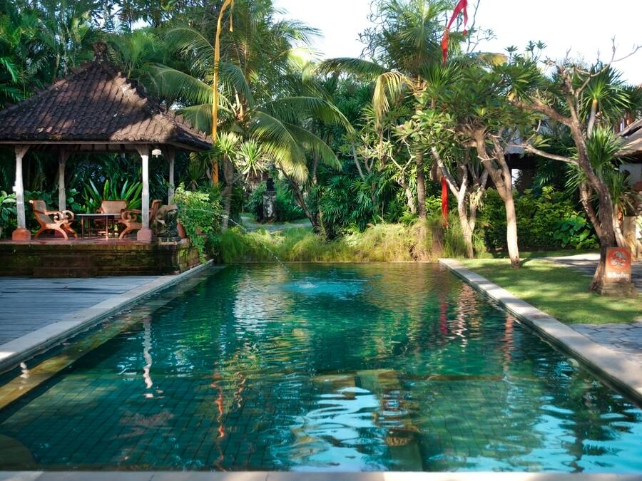 A rectangular pool with clear water, surrounded by lush greenery and a gazebo with chairs.