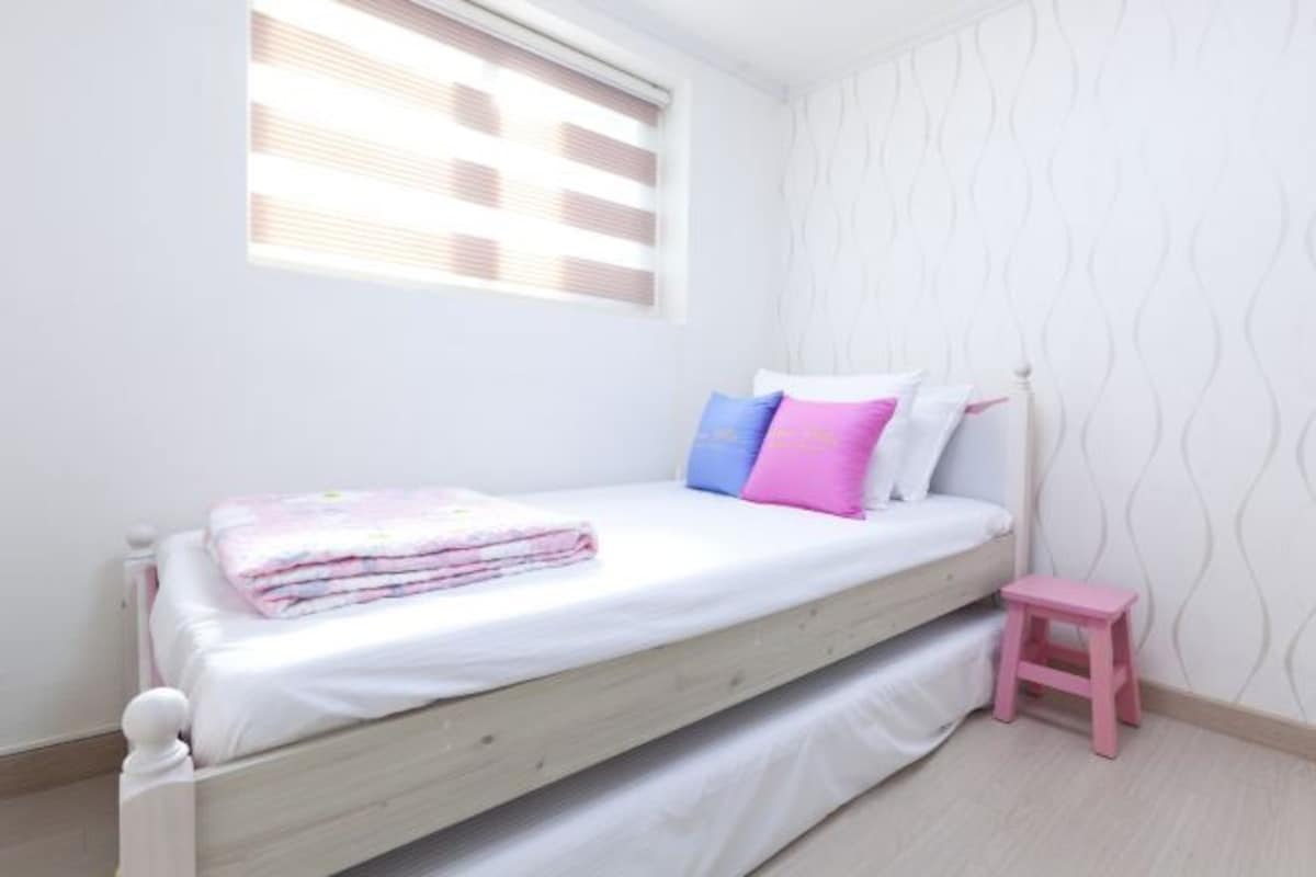 Habitación individual con cama de madera, un taburete pequeño rosa y una ventana con persianas.