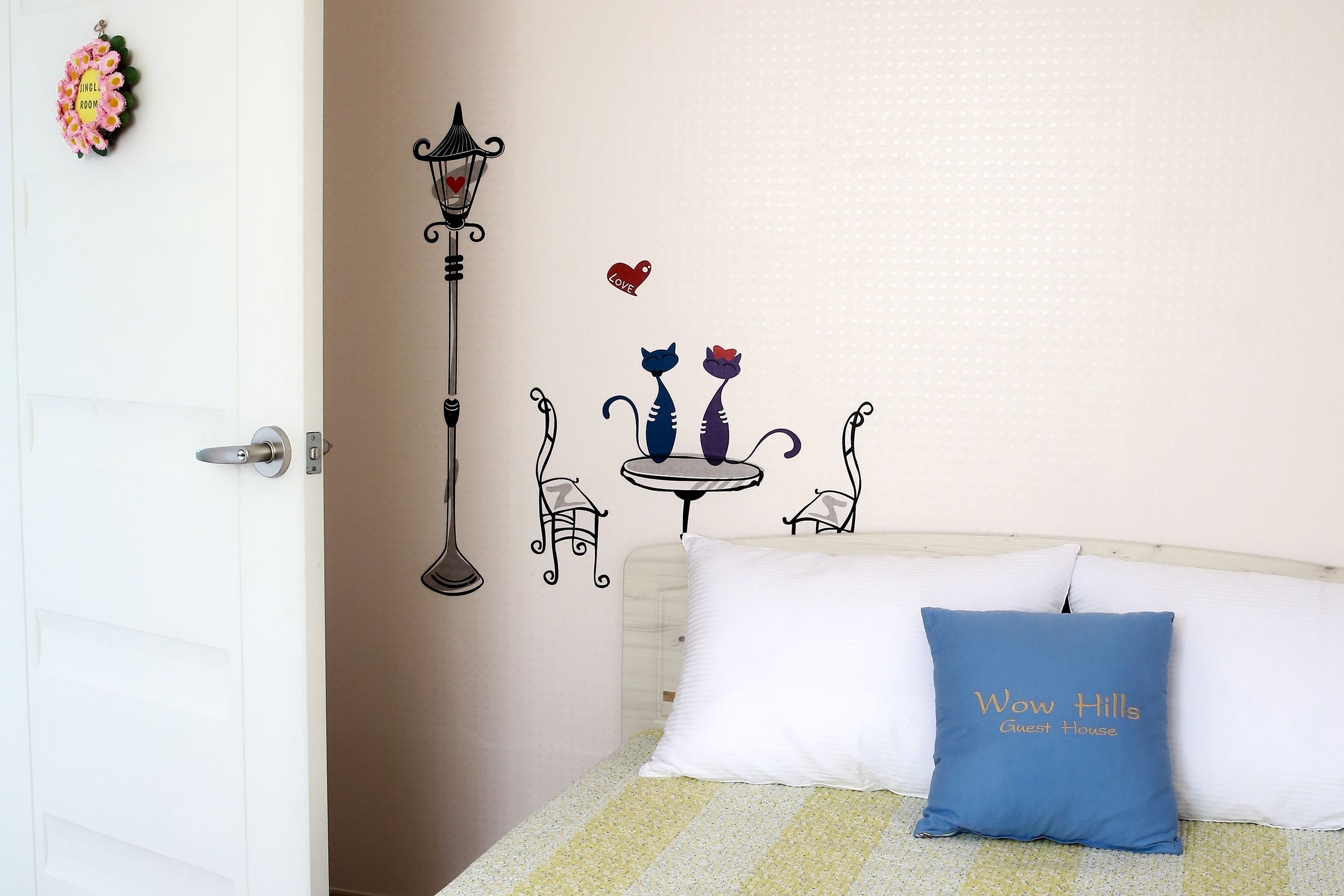 Una camera da letto con un letto, un cuscino blu, una decorazione a parete raffigurante un lampione e un tavolino con due vasi.