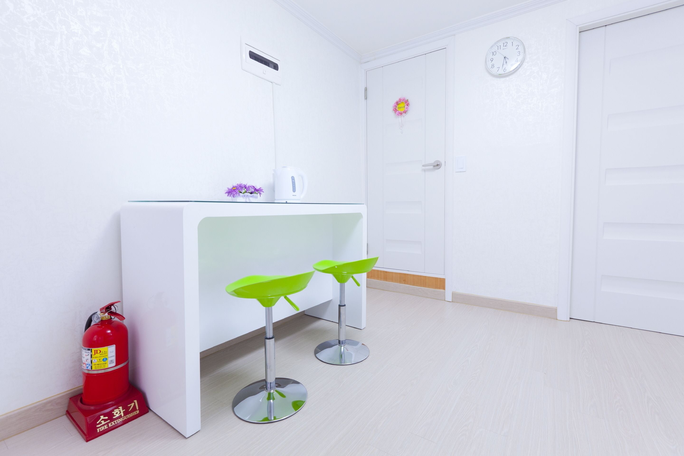A room with a white desk, two green stools, a red fire extinguisher, and a clock on the wall.
