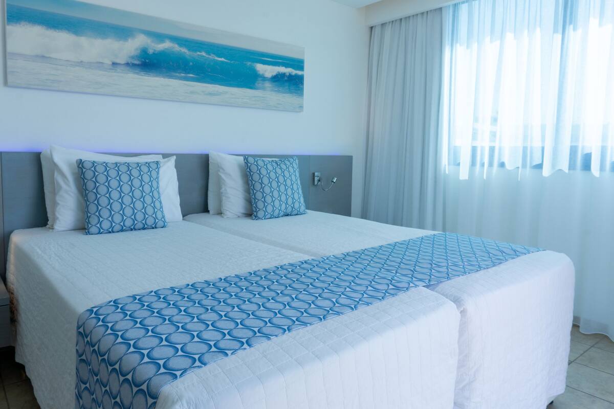 Hotellihuone, jossa on suuri sänky, pääty, meriaiheinen taulu ja ikkuna, jossa on verhot.