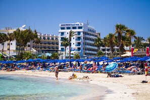 Une plage avec des transats et des parasols, un hôtel appelé «ANONYMOUS Beach Hotel» en arrière-plan, et des palmiers.