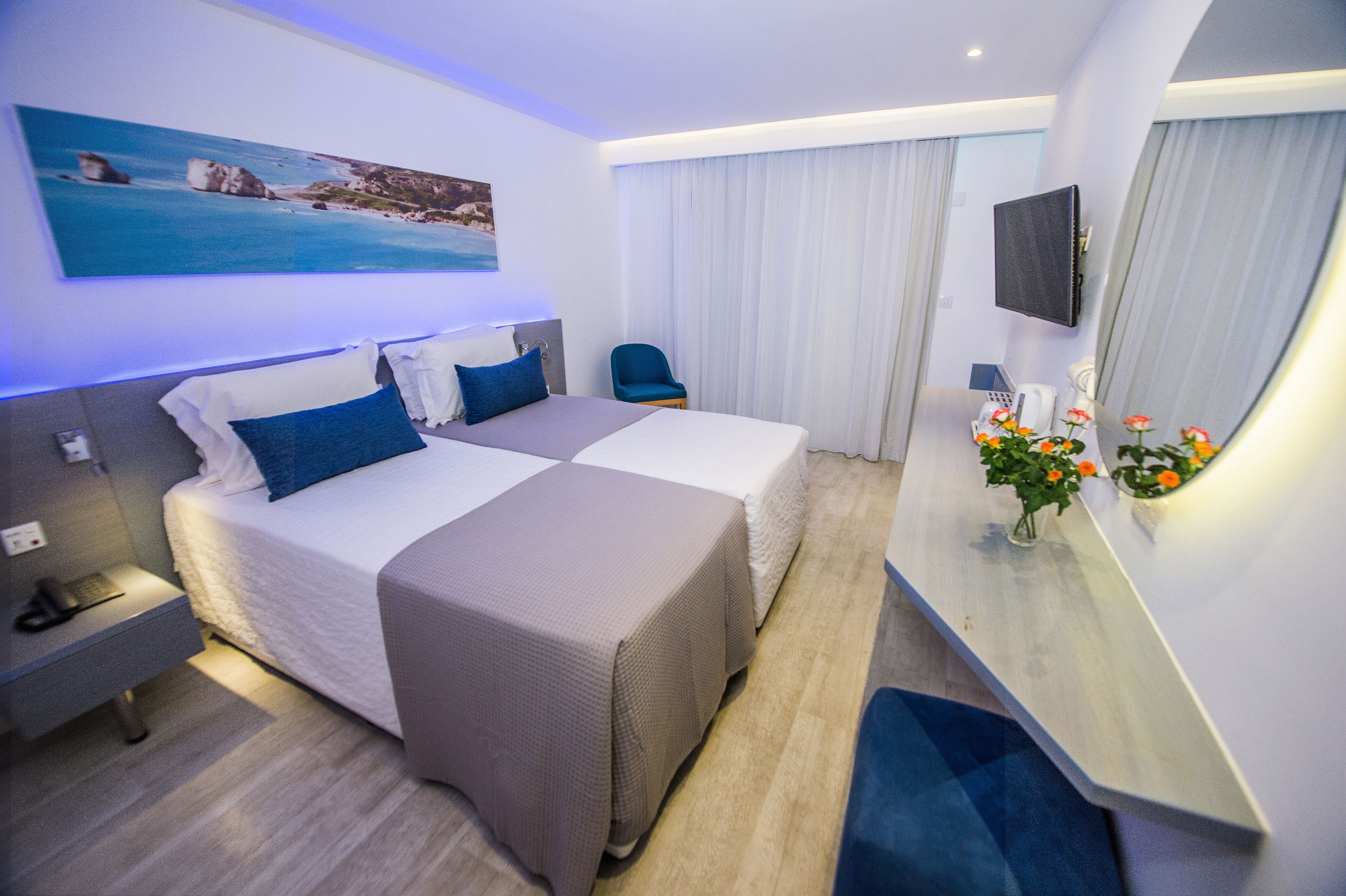 A hotel room with two beds, a desk, a chair, a TV, and a painting of a beach scene.