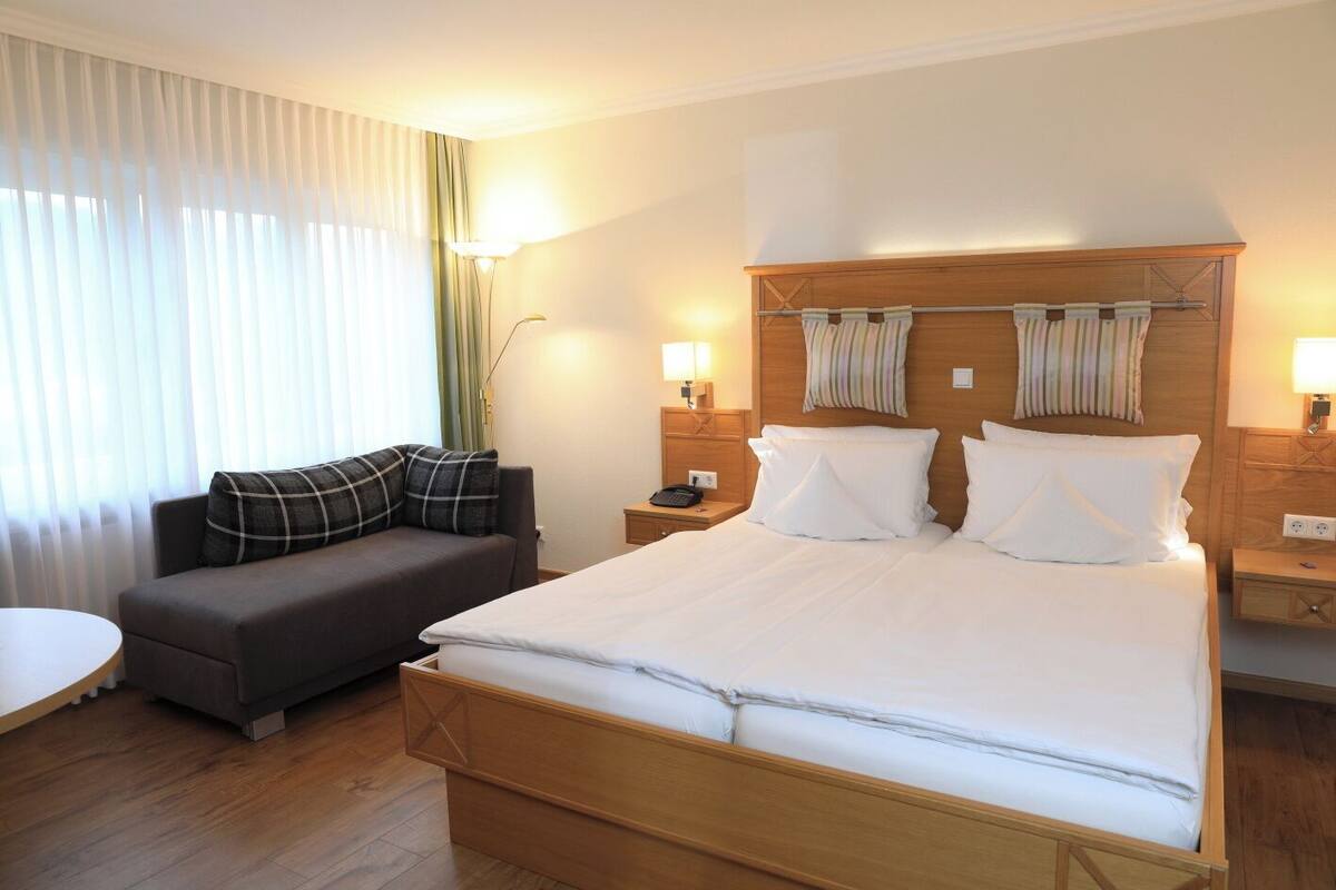 A hotel room with a large bed, a sofa, a bedside table with a lamp, and a window with curtains.