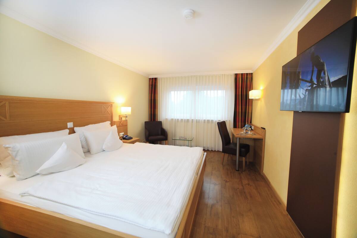 A hotel room with a large bed, a desk with a chair, a television on the wall, and a window with curtains.