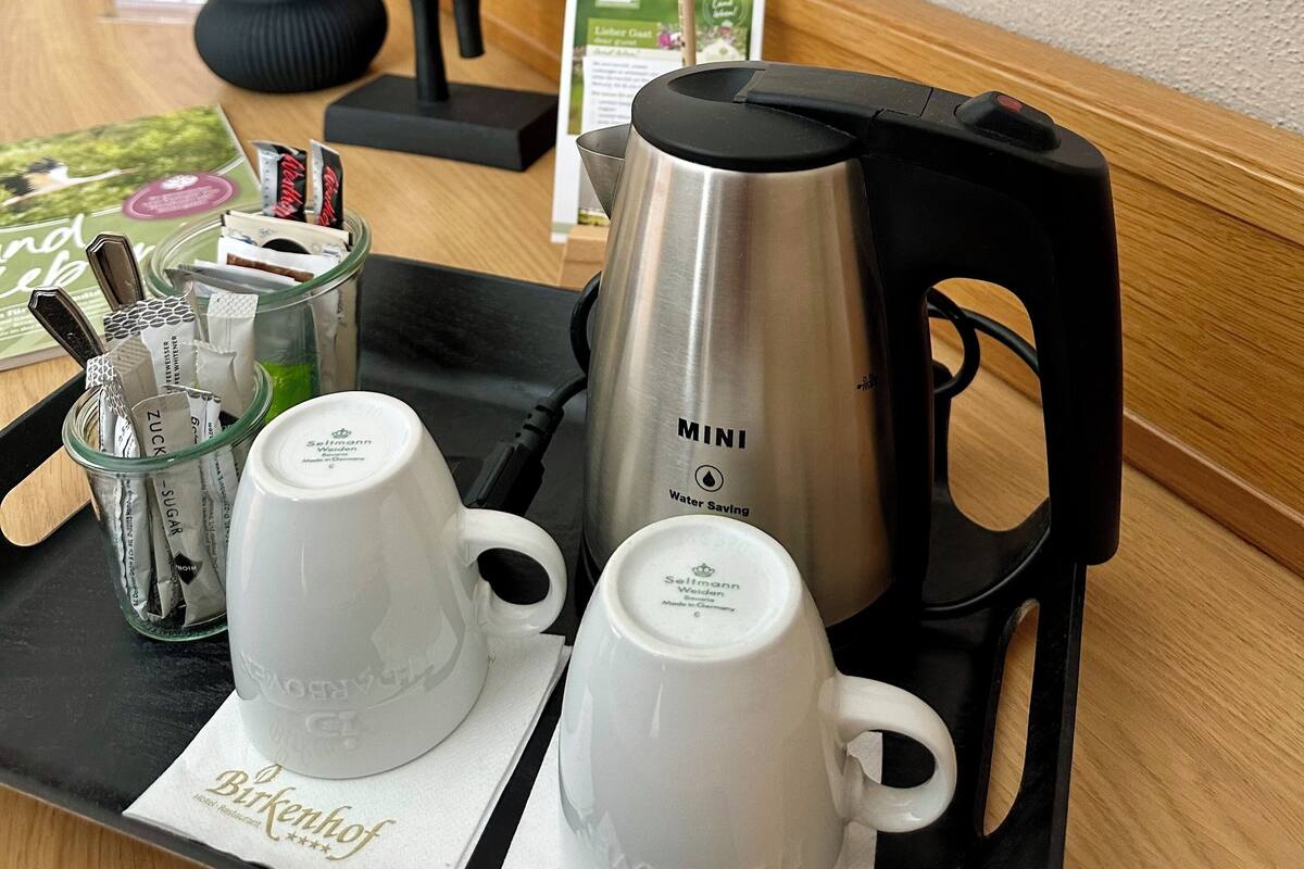 A hotel room with a wooden sideboard, a black abstract sculpture, a vase with dried plants, a tray with a water kettle, two white cups, and a menu.