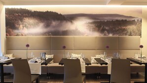 A dining area with tables set for two, each with a vase of purple flowers, glasses, and cutlery. A large wall mural depicts a forest scene with mist. - Landidyll Hotel-Restaurant Birkenhof (Klosterkumbd)