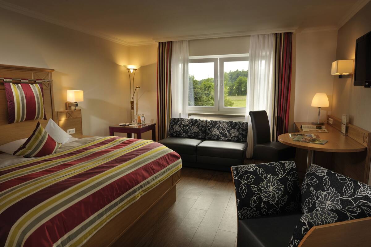 A hotel room with a bed, a sofa, a small table, and a window with curtains.
