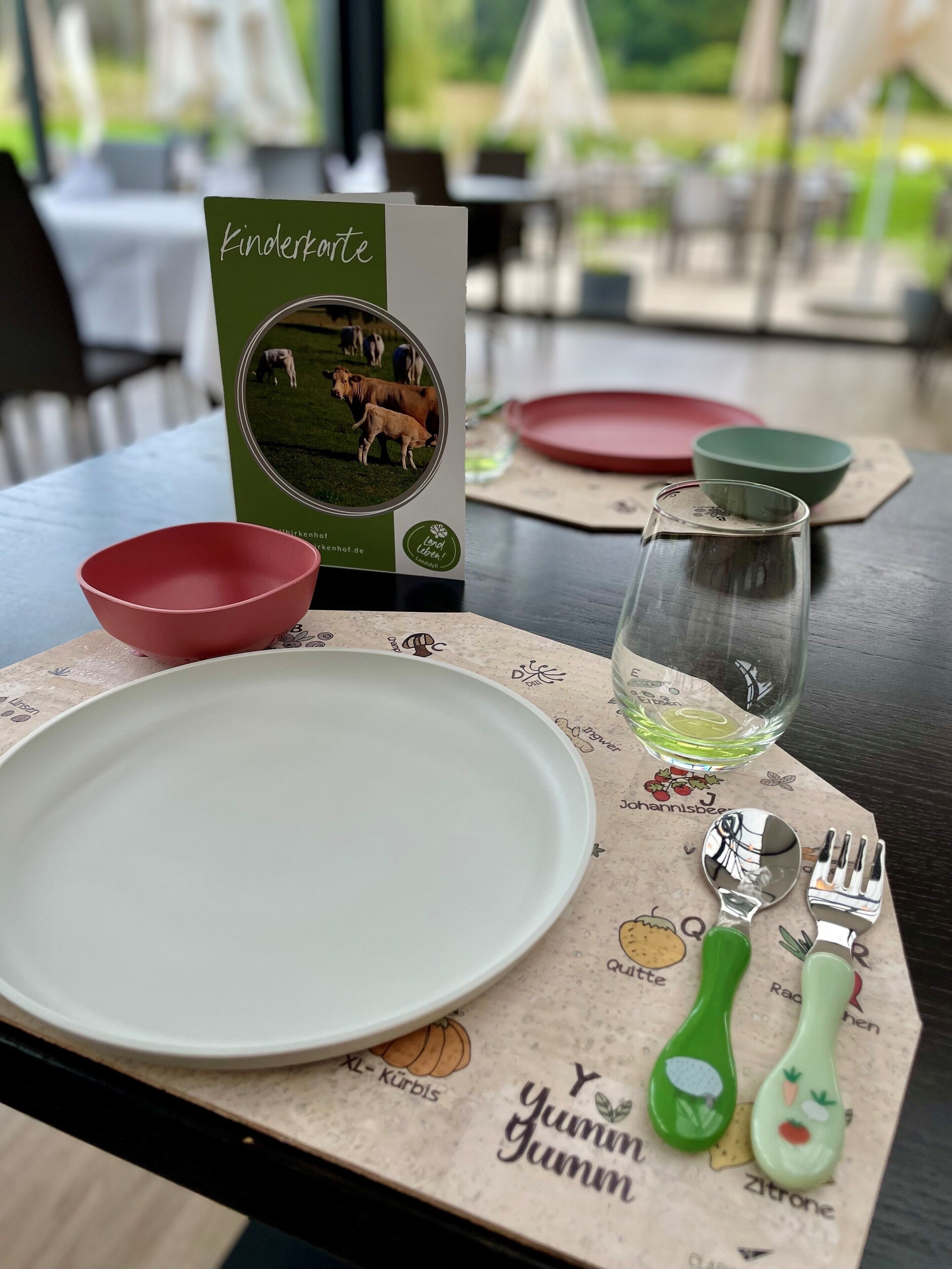 Une table à manger dressée avec une assiette blanche, des bols rouges et verts, un verre d’eau et un menu.