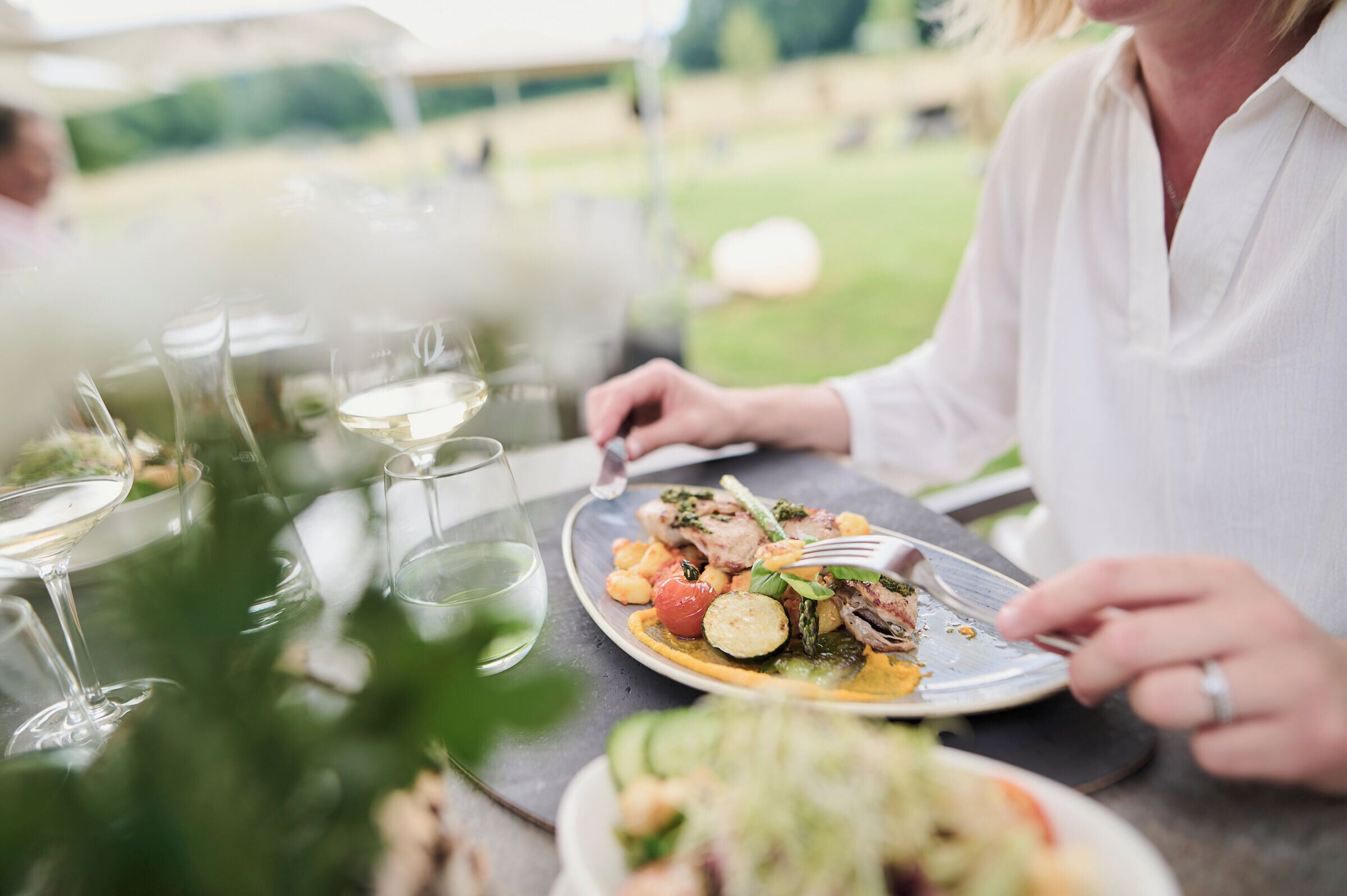 A dining table with plates of food, glasses of white wine, and a person eating.