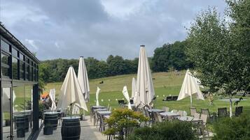 An outdoor seating area with tables and umbrellas, surrounded by greenery and a building with large windows.