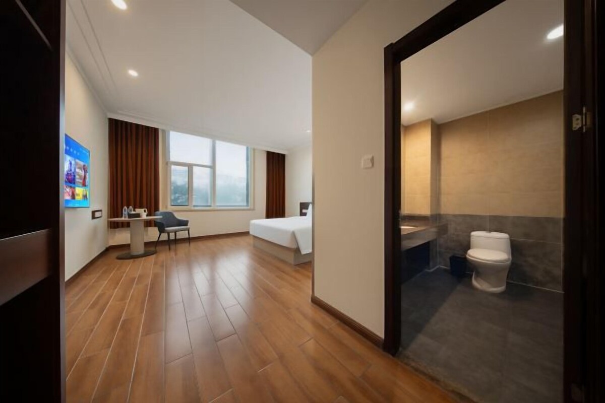 A hotel room with a bed, a desk, a chair, a window, and a bathroom with a toilet and sink.
