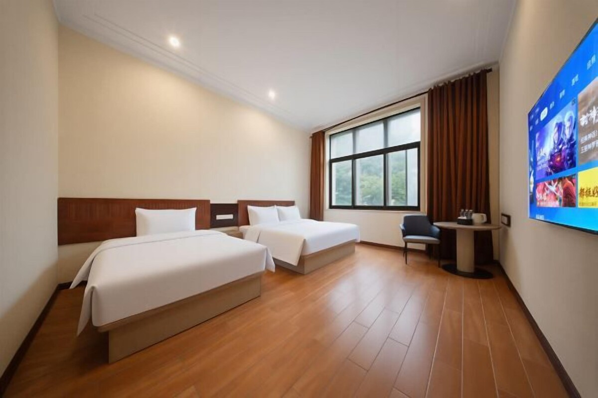 A hotel room with two beds, a desk, a chair, a window, and a wall-mounted screen.