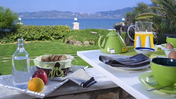 A breakfast table with a view of the sea, including a pitcher of orange juice, a bottle of water, bread, fruit, and a teapot. - Aeolis Apartments & Studios (Lesvos)