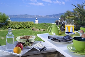 A breakfast table with a view of the sea, including a pitcher of orange juice, a bottle of water, bread, fruit, and a teapot. - Aeolis Apartments & Studios (Lesvos)