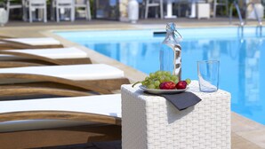 A poolside lounge area with white wicker furniture, a glass bottle, and a plate of fruit. - Aeolis Apartments & Studios (Lesvos)