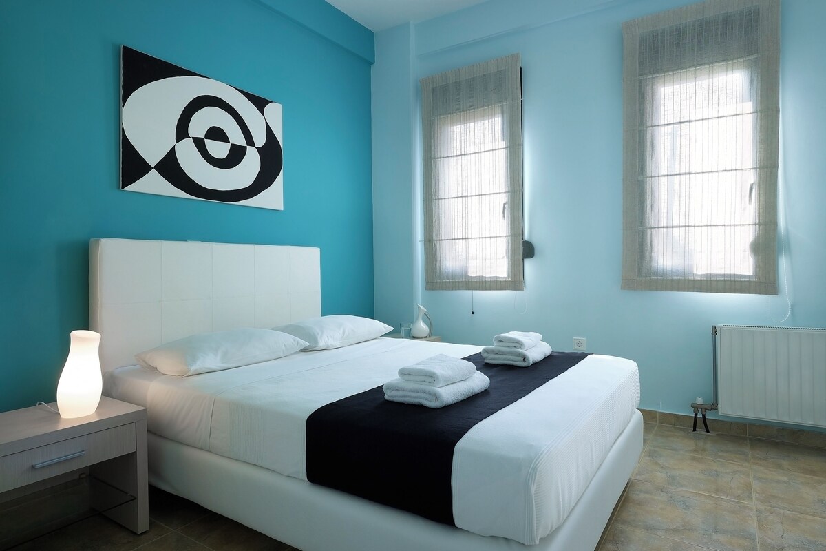 Une chambre moderne avec un grand lit, une table de chevet avec une lampe et deux fenêtres équipées de stores.