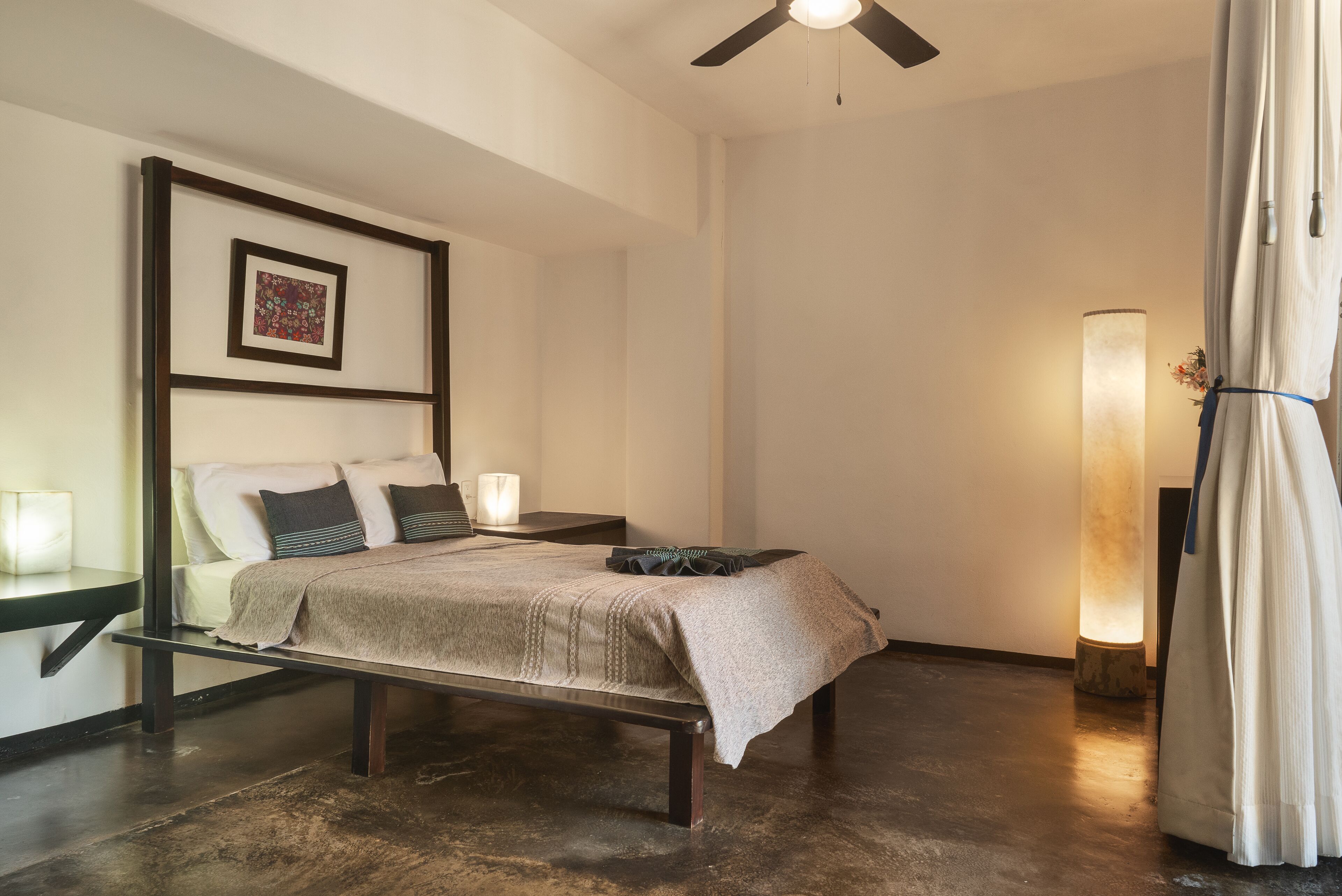 A bedroom with a bed, bedside tables, a ceiling fan, a wall-mounted artwork, and a floor lamp.