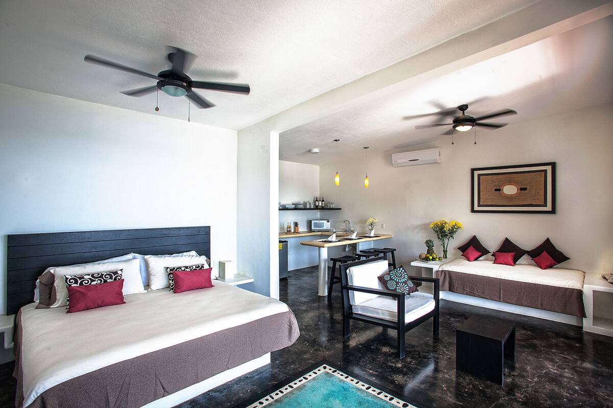 A hotel room with a bed, a desk, a chair, and a kitchenette in the background.