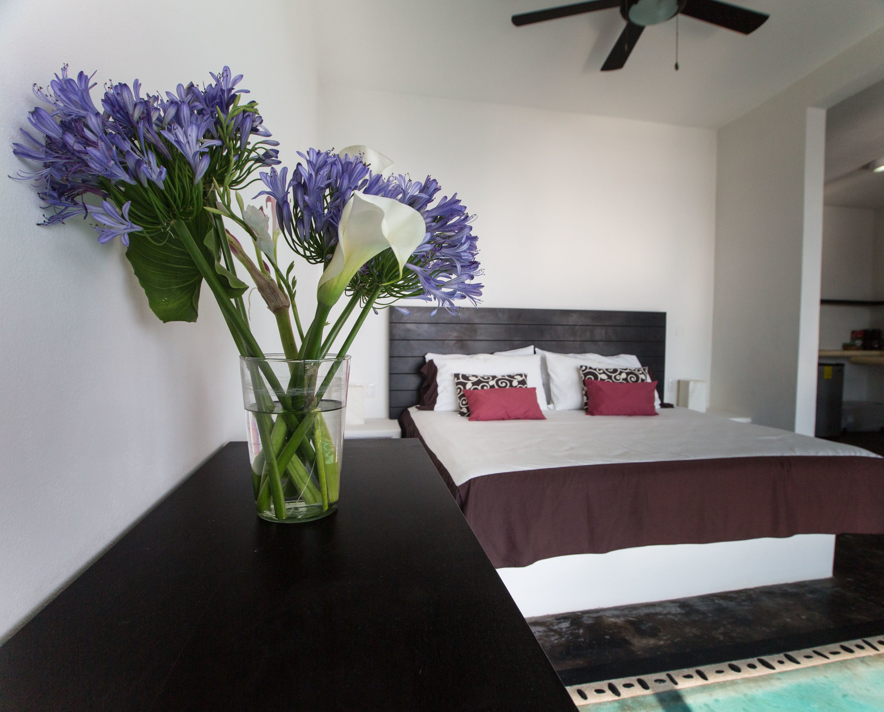 Een slaapkamer met een bed, kussens, een nachtkastje en een vaas met bloemen.