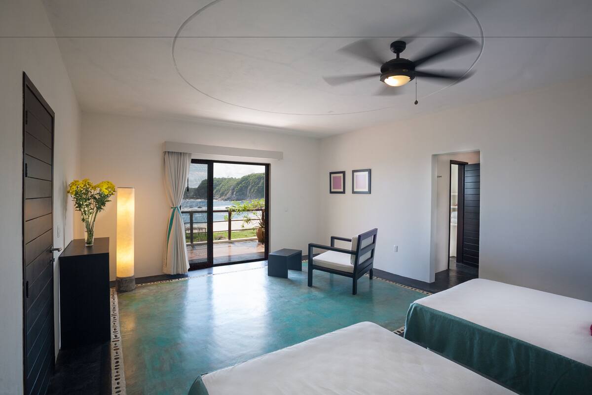 A hotel room with a bed, a chair, a desk, a ceiling fan, and a view of the outdoors.