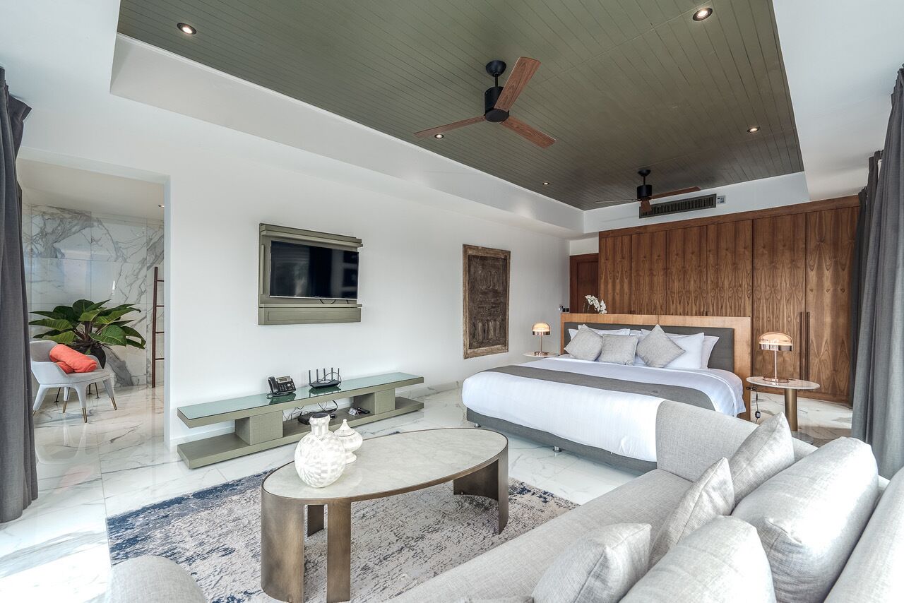 Un dormitorio moderno con una cama grande, un televisor montado en la pared, un ventilador de techo de madera y una zona de estar con una mesa de centro.