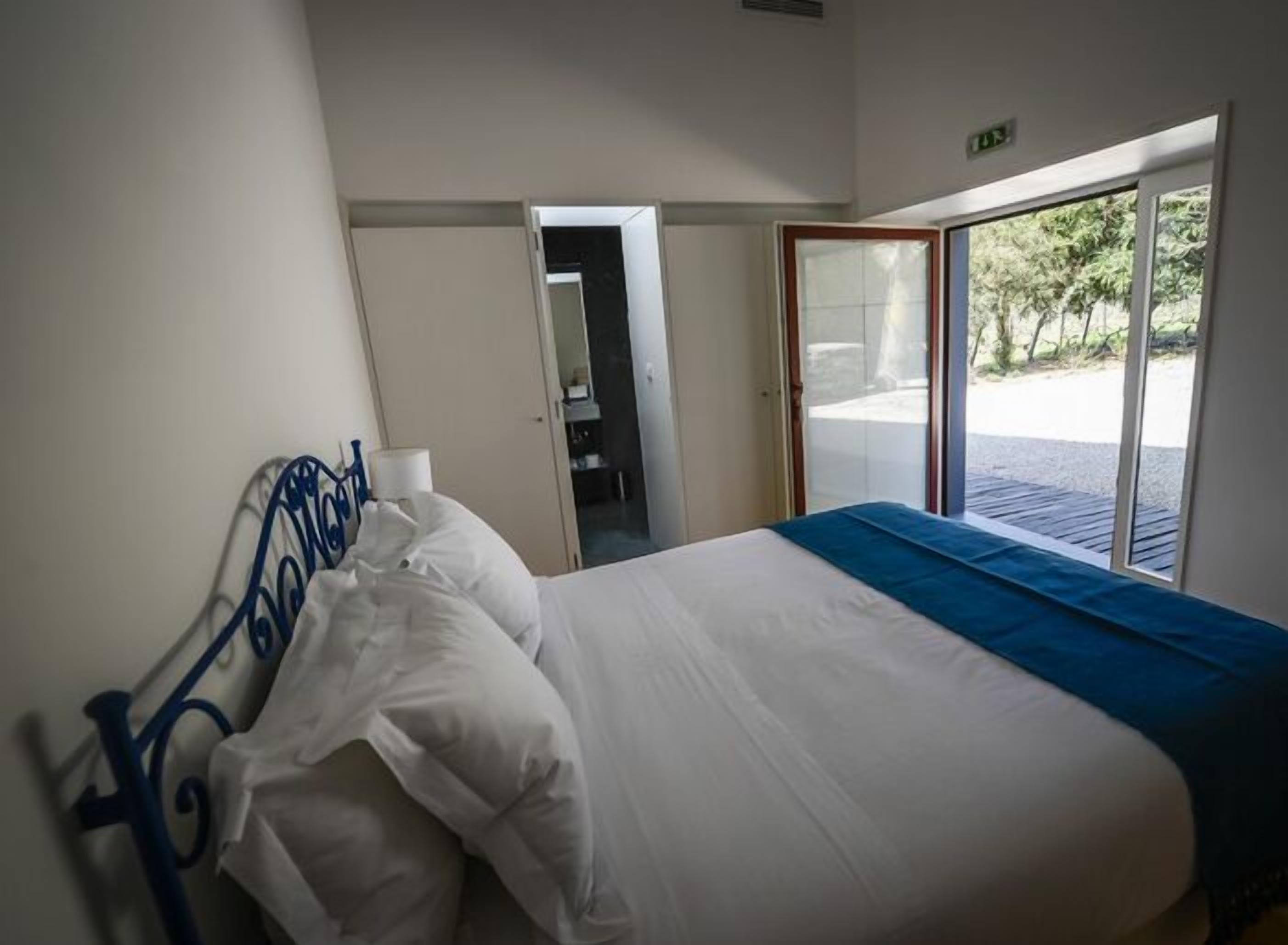 A hotel room with a bed, a window with a view of greenery, and a door leading to another room.