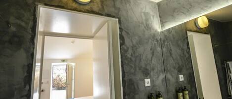 A modern bathroom with a large mirror, a bathtub, and two sinks. There are toiletries on the wall and a door leading to another room.