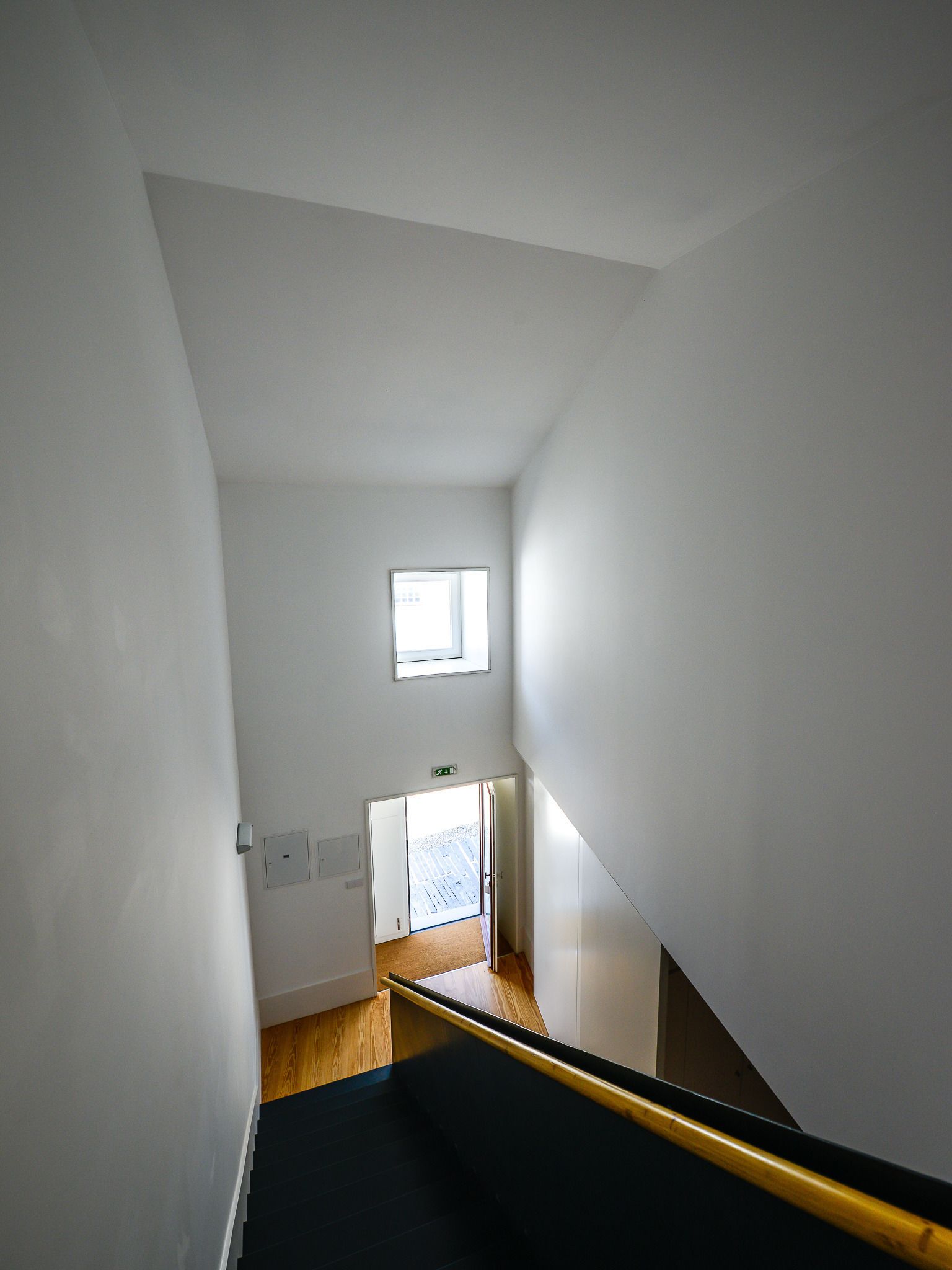 A narrow stairway with a black handrail and wooden flooring, leading to a door with a small window above it.
