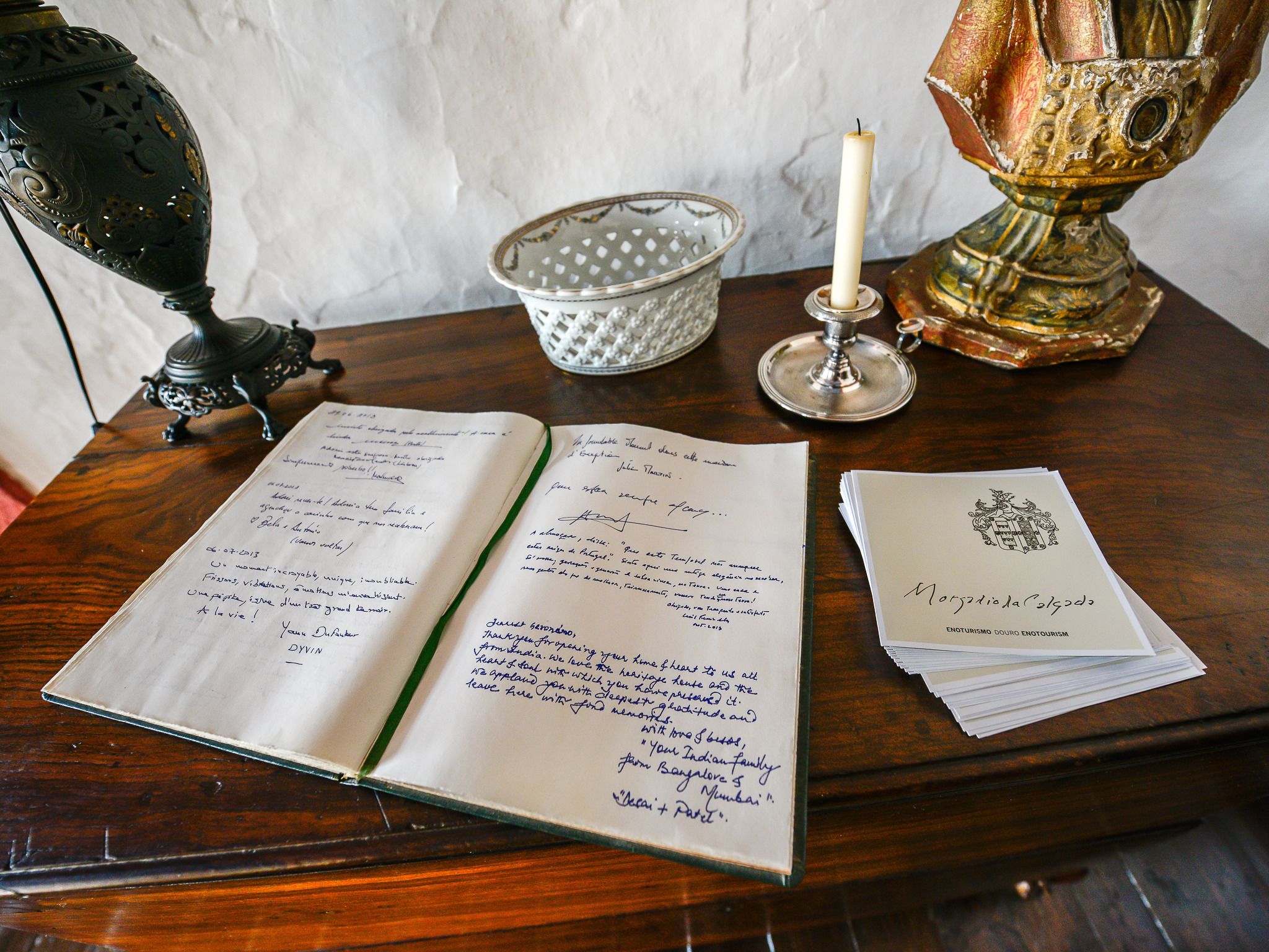 An open notebook with handwritten notes, a candle, a decorative lamp, and a stack of papers on a wooden table.