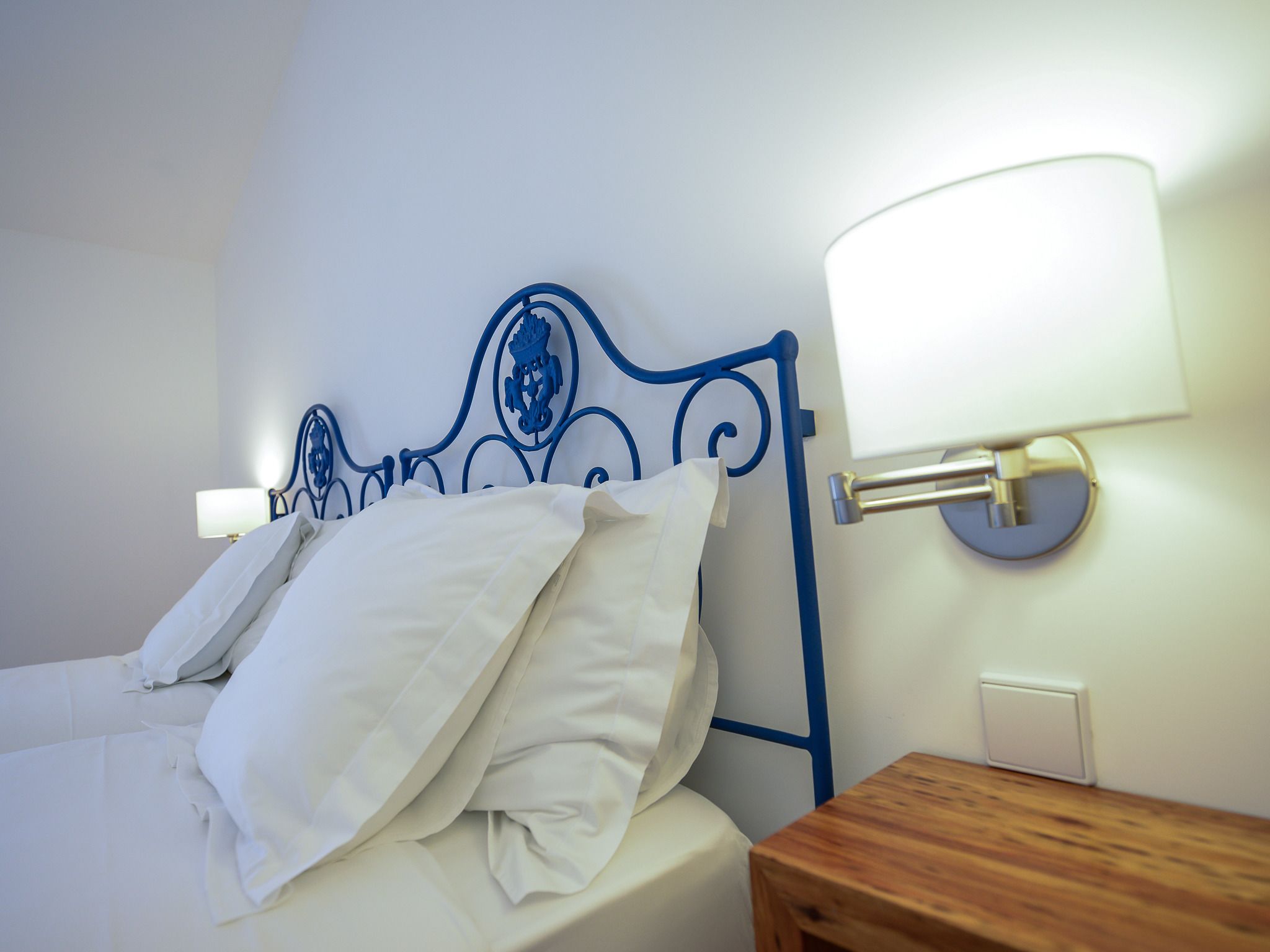 A neatly made bed with white linens and pillows, a blue metal headboard, and two wall-mounted lamps on either side.