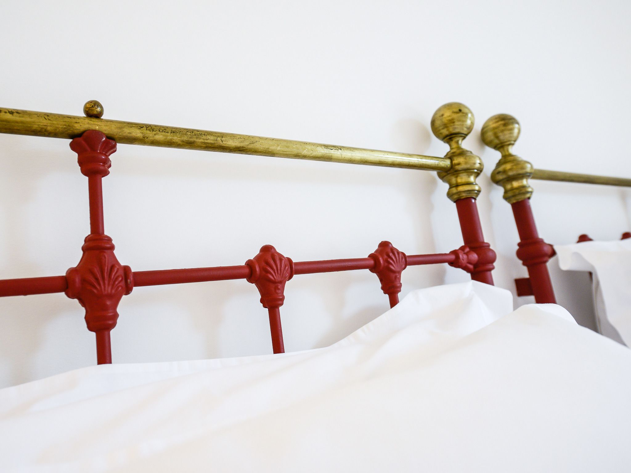 A bed with a red metal frame and brass accents, white bedding, and a white wall in the background.