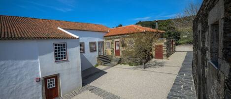 A narrow street with white buildings and red-tiled roofs, stone walls, and a gravel path.