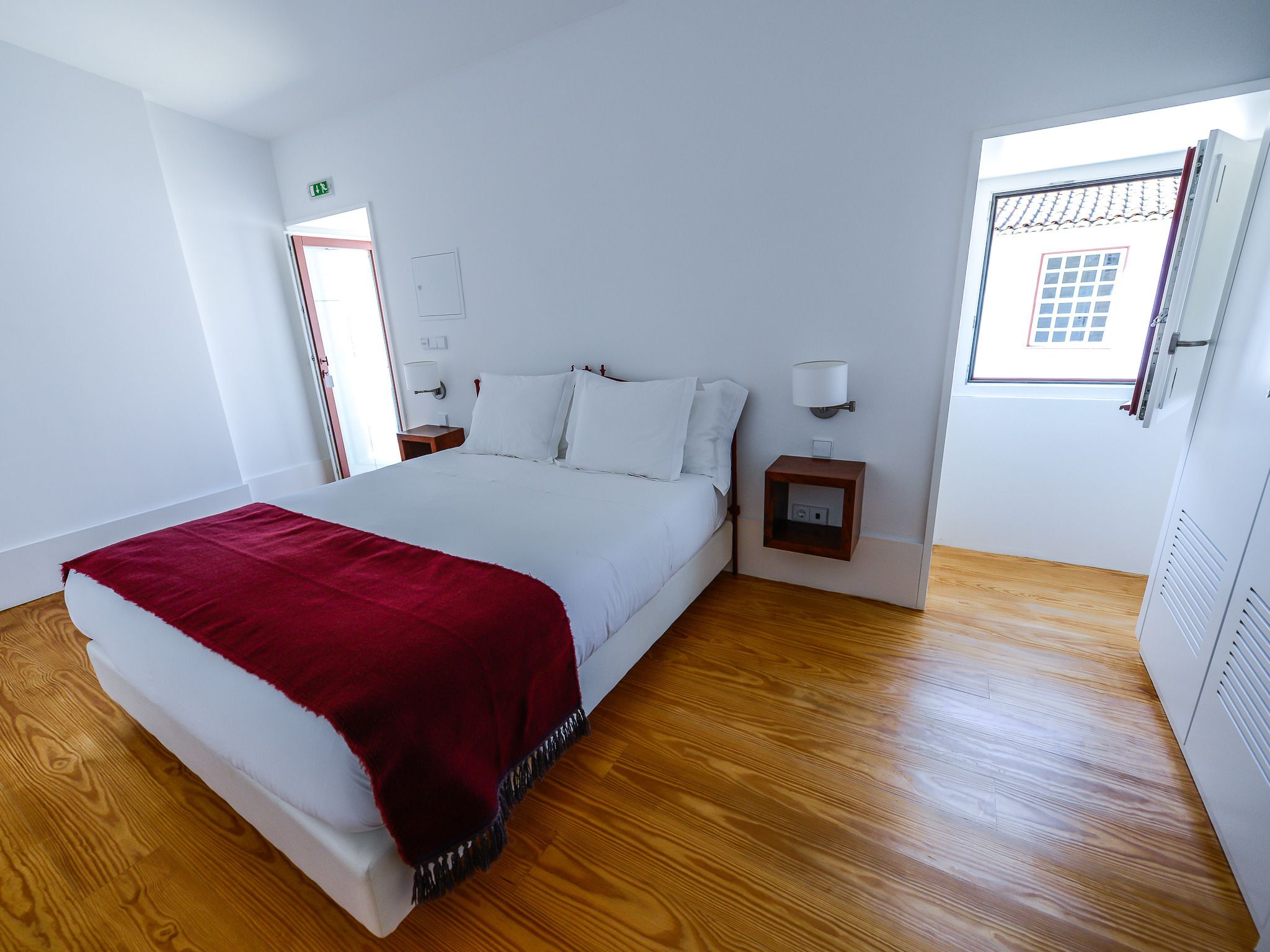 Habitación de hotel con una cama grande, suelo de madera, una mesita de noche con lámpara, una ventana con vista a un edificio y una puerta con letrero de salida de emergencia.