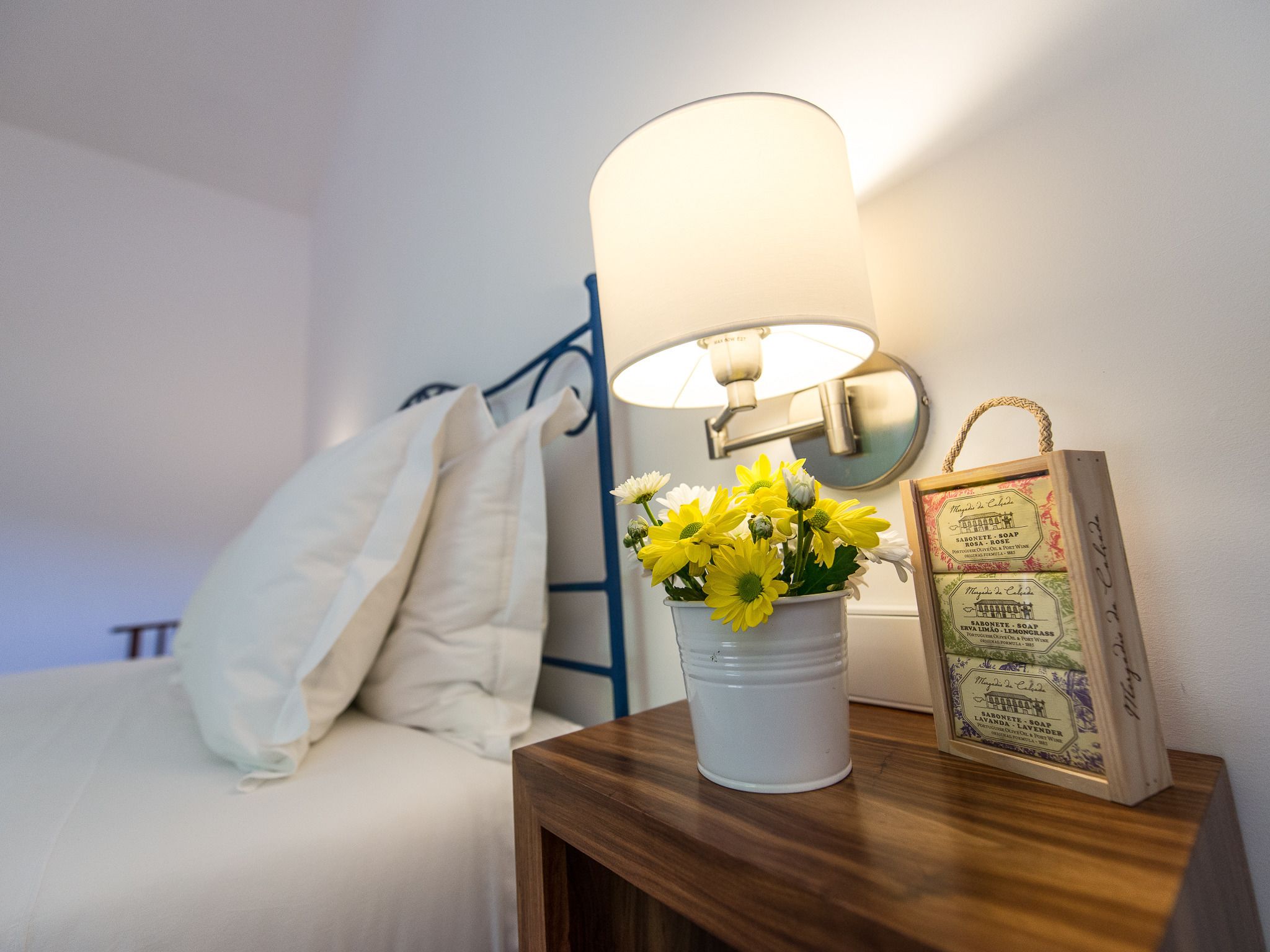 A bedside table with a white vase of yellow flowers, a framed menu, and a wall-mounted lamp.