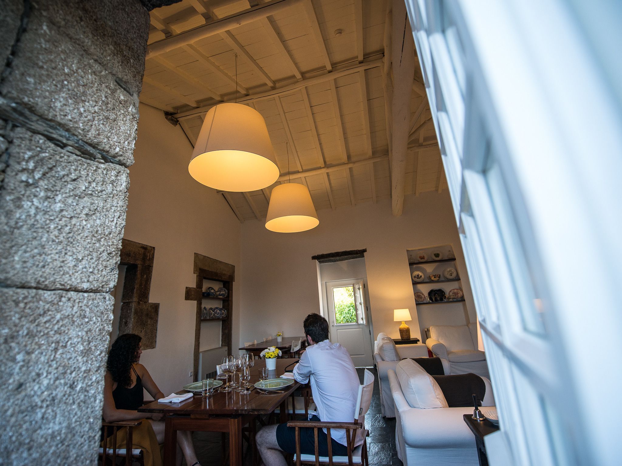 A dining area with a wooden table set for two, a stone wall, and a window with a view of greenery.