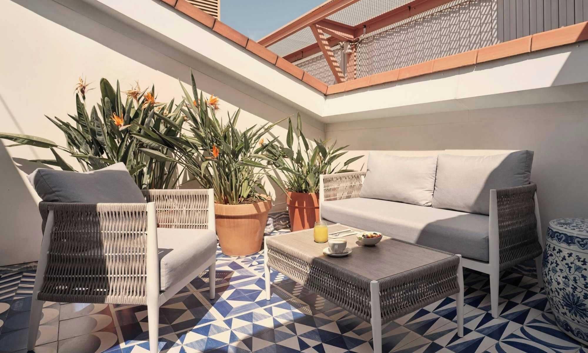 Un espacio al aire libre iluminado por el sol con un sofá, dos sillones, una mesita y plantas en macetas.
