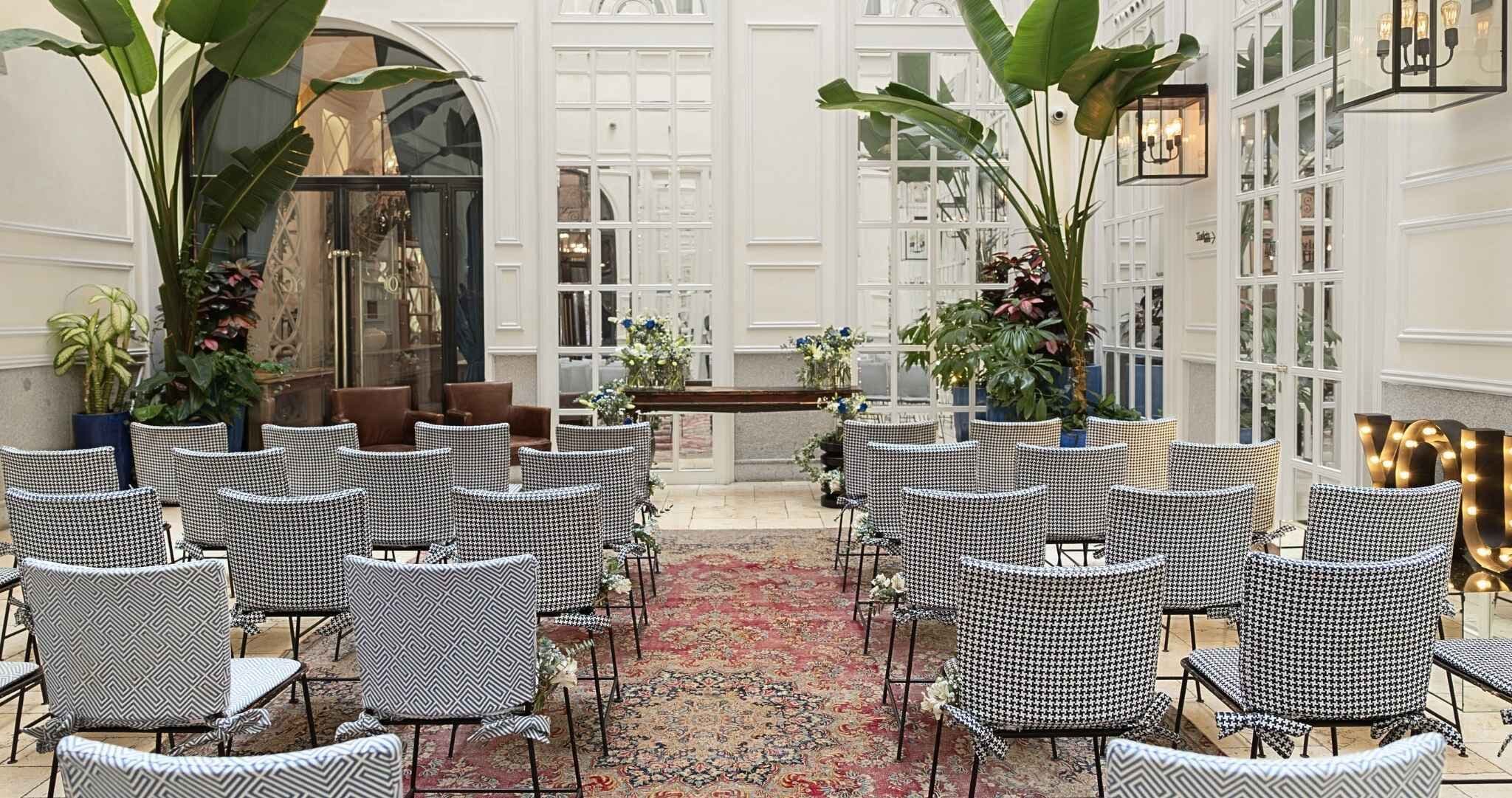 A formal indoor setting with rows of checkered chairs, a central aisle, and potted plants.